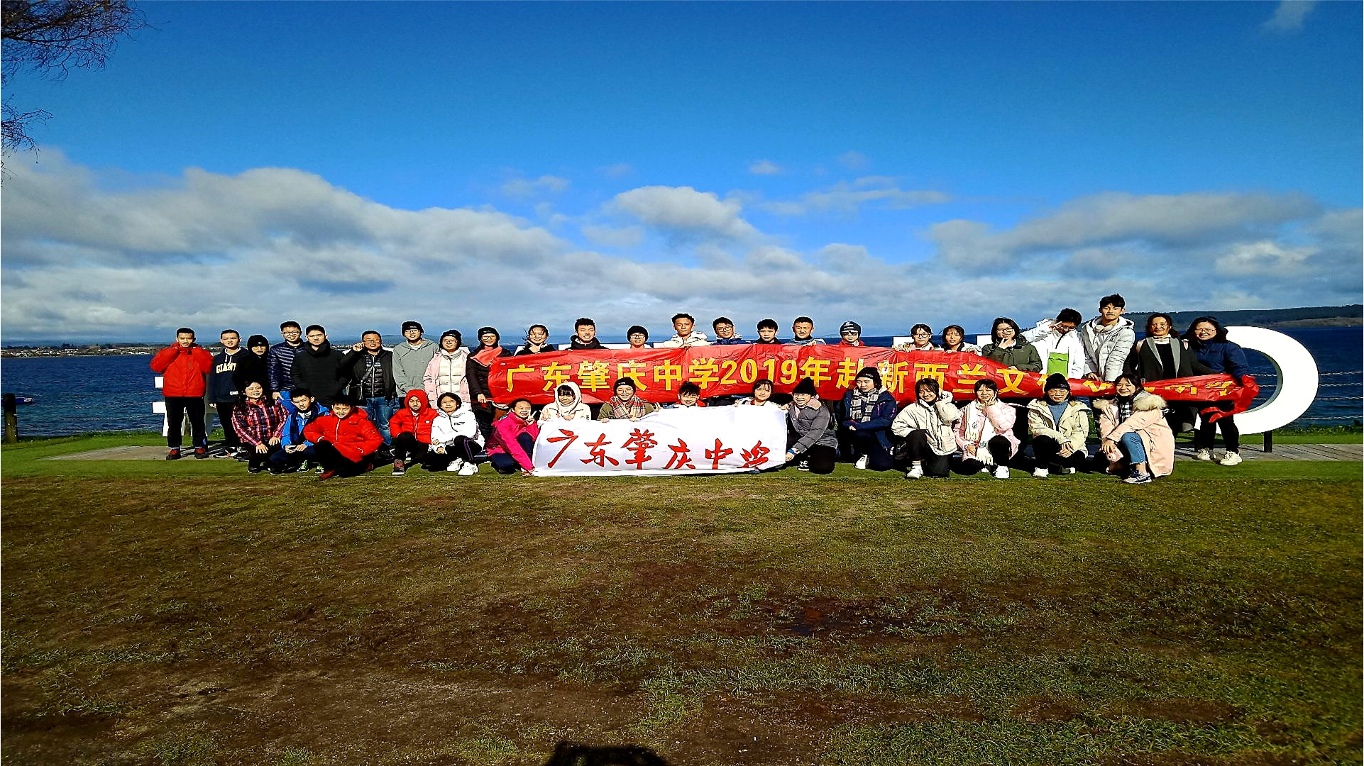 A study tour group posing with a banner on a grassy lakeside